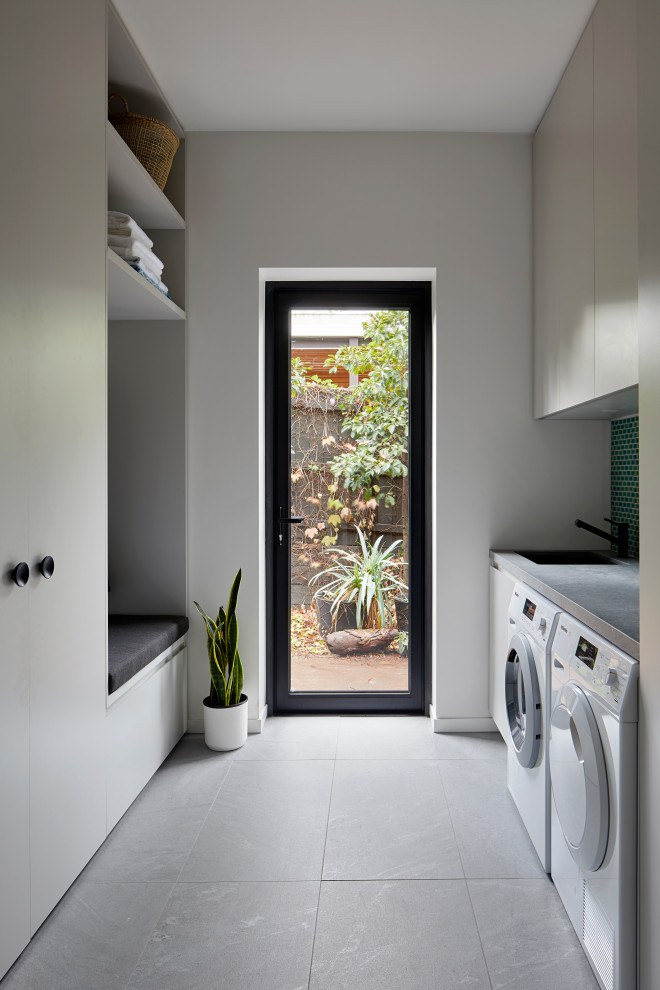 Exempel på en stor modern grå parallell grått tvättstuga enbart för tvätt, med en nedsänkt diskho, släta luckor, vita skåp, grönt stänkskydd, stänkskydd i mosaik, vita väggar, klinkergolv i porslin, en tvättmaskin och torktumlare bredvid varandra och grått golv