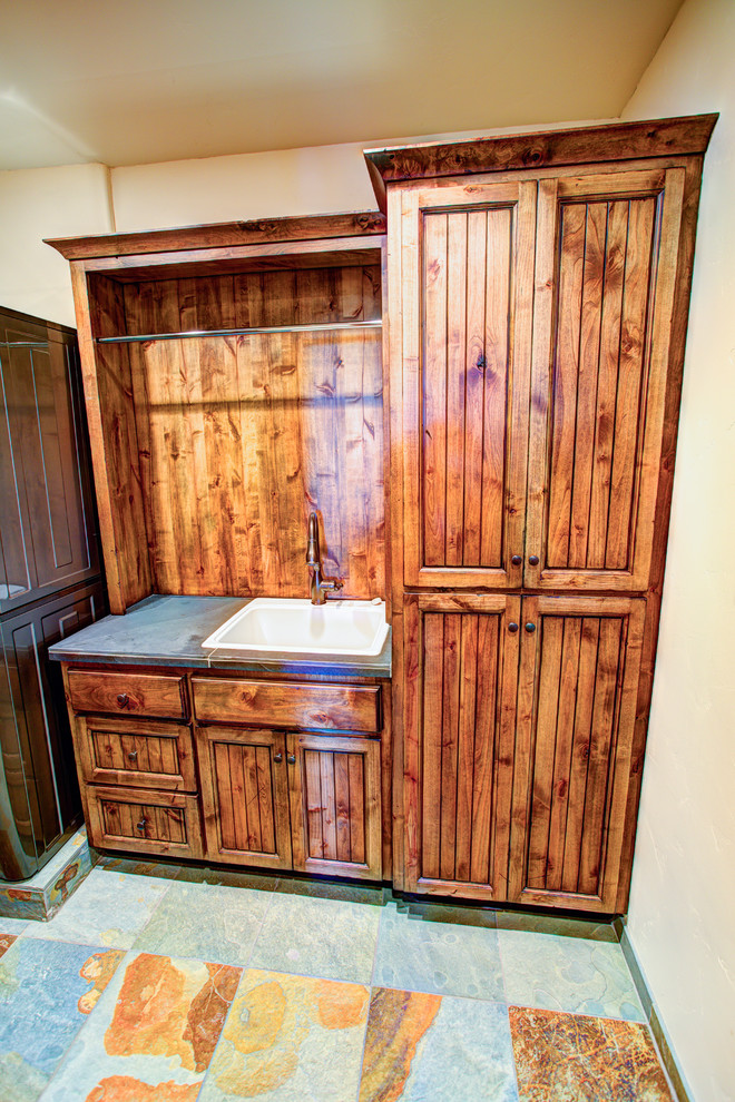 Inredning av en rustik mellanstor linjär tvättstuga enbart för tvätt, med luckor med infälld panel, bänkskiva i kalksten, beige väggar, skiffergolv, en tvättpelare, skåp i mörkt trä och en nedsänkt diskho