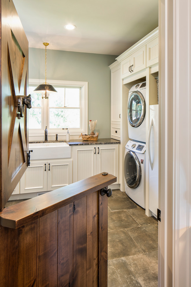 Inspiration for a large farmhouse l-shaped dedicated laundry room remodel in Minneapolis with a farmhouse sink, recessed-panel cabinets, white cabinets, granite countertops, a stacked washer/dryer and gray walls
