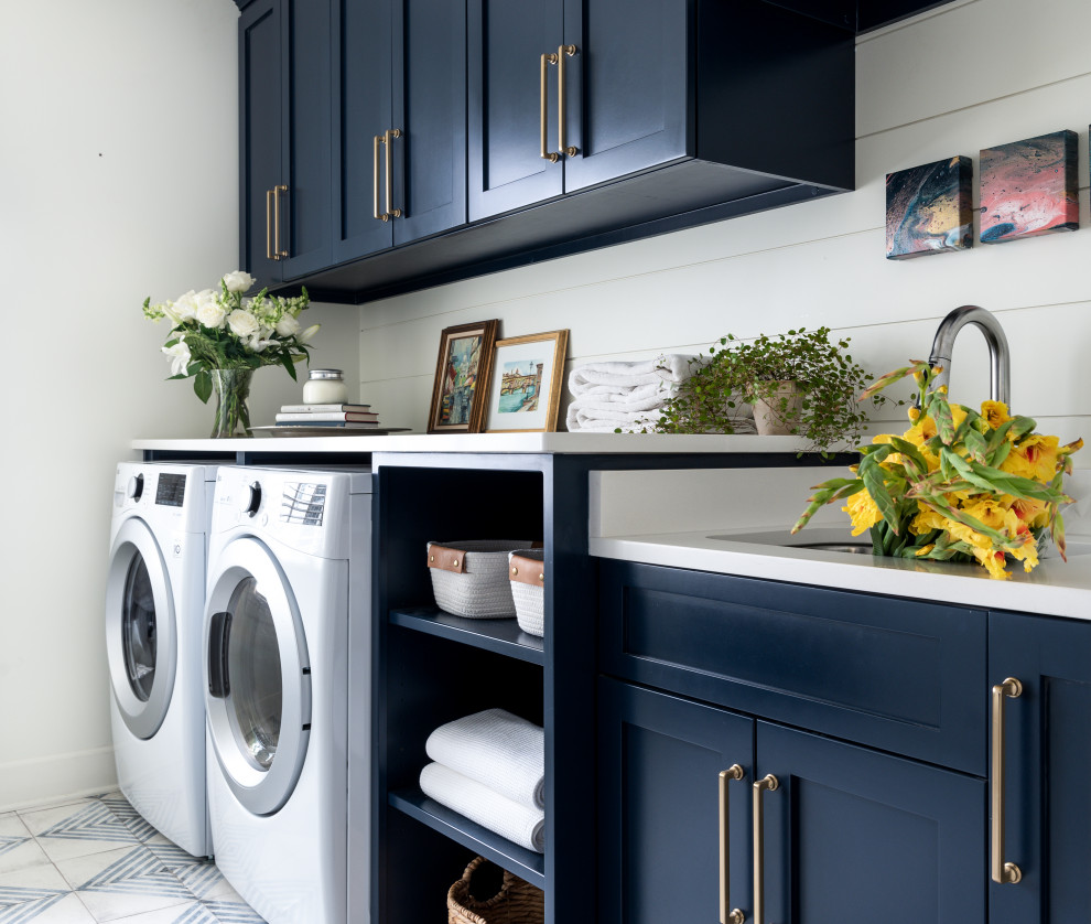 ナッシュビルにあるお手頃価格の広いトランジショナルスタイルのおしゃれな洗濯室 (ll型、アンダーカウンターシンク、シェーカースタイル扉のキャビネット、青いキャビネット、クオーツストーンカウンター、塗装板のキッチンパネル、白い壁、磁器タイルの床、左右配置の洗濯機・乾燥機、マルチカラーの床、白いキッチンカウンター、塗装板張りの壁) の写真