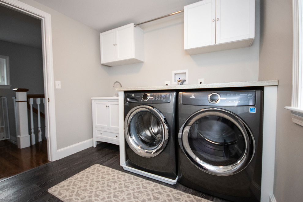 Klassisk inredning av en mellanstor bruna linjär brunt tvättstuga enbart för tvätt, med en nedsänkt diskho, luckor med infälld panel, vita skåp, laminatbänkskiva, grå väggar, vinylgolv, en tvättmaskin och torktumlare bredvid varandra och grått golv