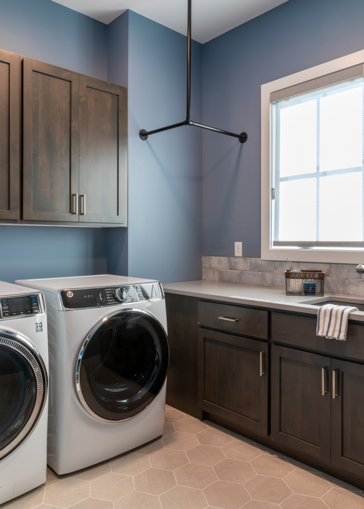 Bild på en stor industriell grå l-formad grått tvättstuga enbart för tvätt, med en undermonterad diskho, skåp i shakerstil, grå skåp, bänkskiva i kvartsit, blått stänkskydd, stänkskydd i keramik, blå väggar, klinkergolv i keramik, en tvättmaskin och torktumlare bredvid varandra och grått golv