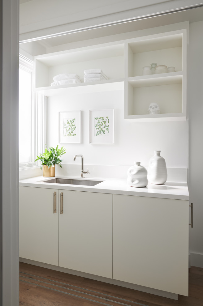Photo of a large traditional single-wall laundry cupboard in San Francisco with a submerged sink, flat-panel cabinets, white cabinets, engineered stone countertops, white walls, light hardwood flooring, a stacked washer and dryer and white worktops.