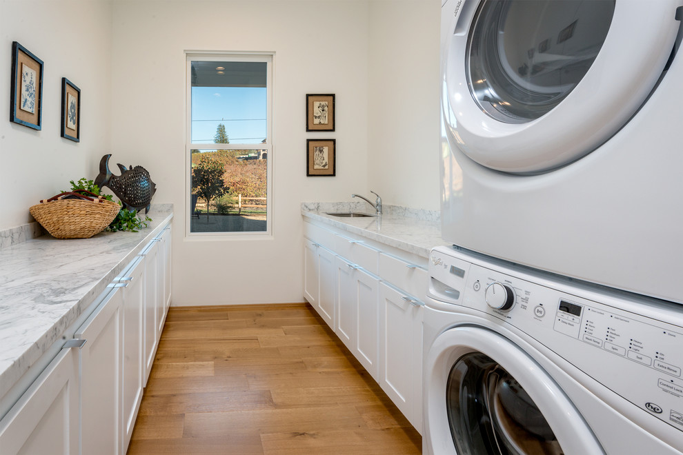 Farmhouse galley light wood floor dedicated laundry room photo in San Francisco with an undermount sink, shaker cabinets, white cabinets, marble countertops, white walls and a stacked washer/dryer
