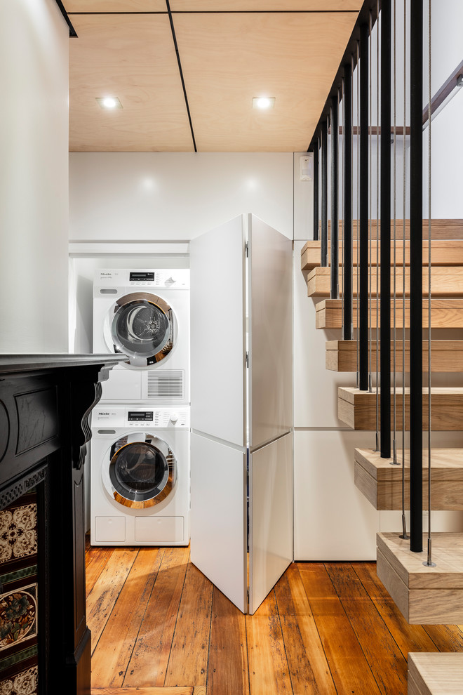 Einzeiliger Moderner Hauswirtschaftsraum mit Waschmaschinenschrank, flächenbündigen Schrankfronten, weißen Schränken, weißer Wandfarbe, braunem Holzboden, Waschmaschine und Trockner gestapelt und braunem Boden in Sydney