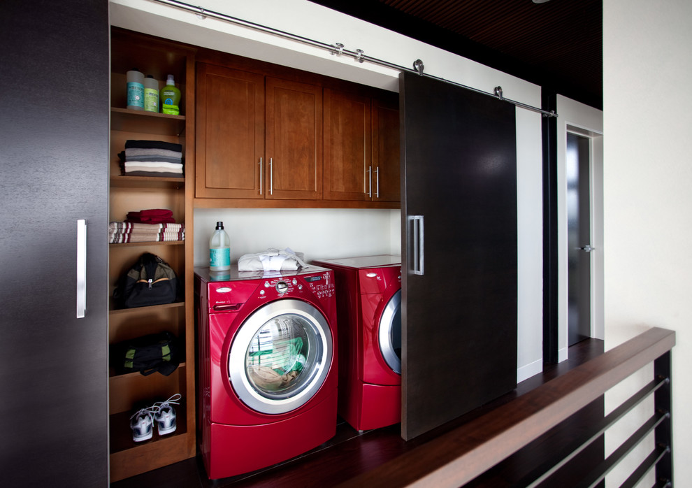 Diseño de armario lavadero lineal contemporáneo pequeño con armarios con paneles empotrados, puertas de armario de madera oscura, paredes blancas, suelo de madera oscura y lavadora y secadora juntas