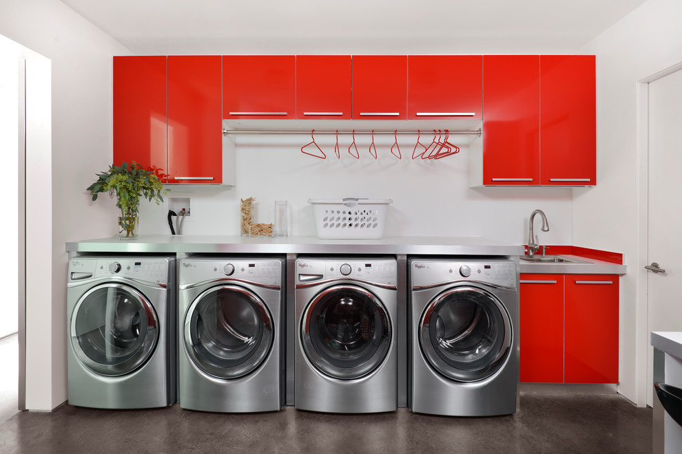 Modelo de lavadero multiusos actual con pila para lavar, puertas de armario rojas, paredes blancas, suelo de cemento, lavadora y secadora juntas y suelo gris