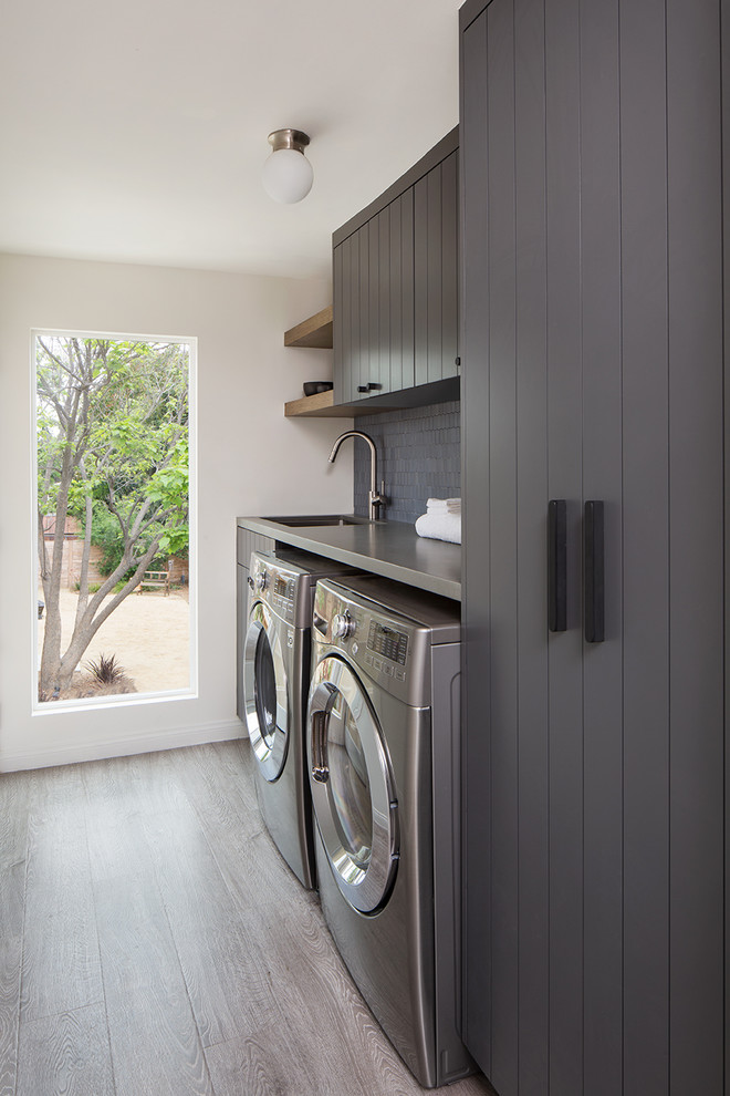 Foto på en liten funkis grå tvättstuga enbart för tvätt, med grå skåp, bänkskiva i kvarts, vita väggar, ljust trägolv, en tvättmaskin och torktumlare bredvid varandra, grått golv och en undermonterad diskho