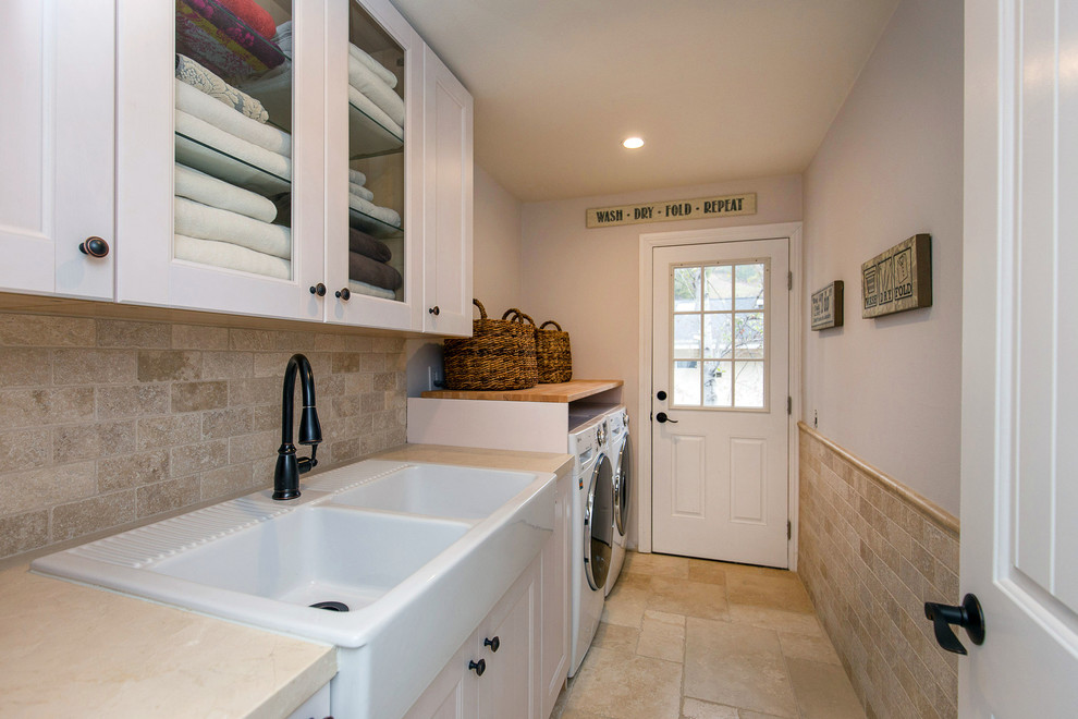 Example of a small farmhouse galley travertine floor dedicated laundry room design in Los Angeles with a farmhouse sink, shaker cabinets, white cabinets, wood countertops, gray walls, a side-by-side washer/dryer and beige countertops