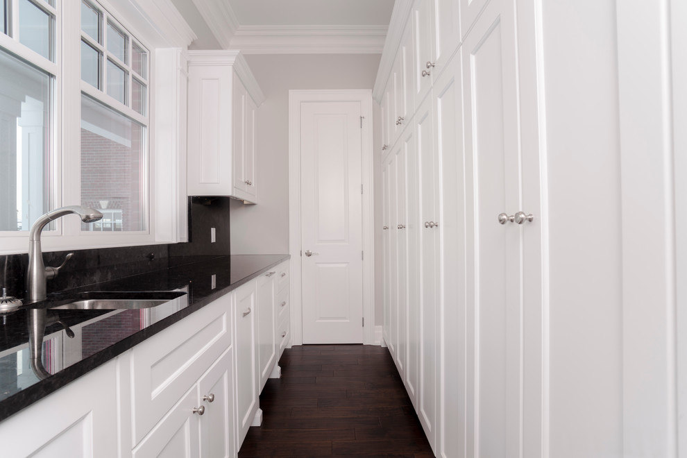 Ejemplo de cuarto de lavado lineal tradicional con fregadero encastrado, puertas de armario blancas, paredes blancas, suelo de madera oscura, lavadora y secadora apiladas y armarios con paneles empotrados
