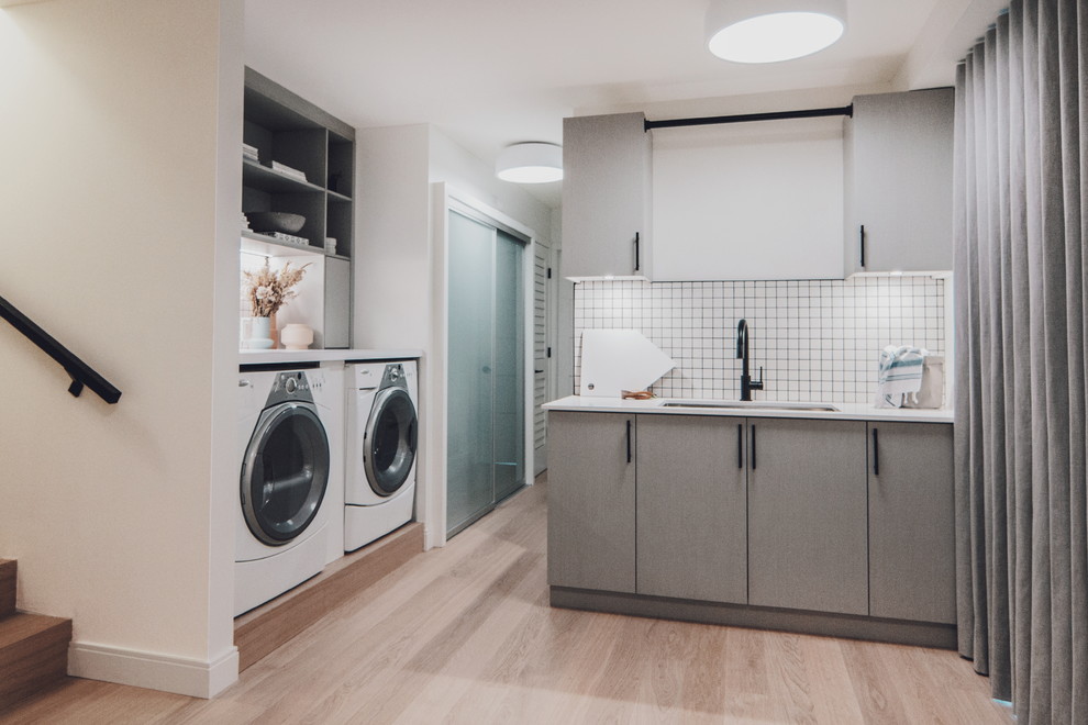 Inspiration for a contemporary utility room in Vancouver with a submerged sink, flat-panel cabinets, grey cabinets, engineered stone countertops, white walls, light hardwood flooring and a side by side washer and dryer.