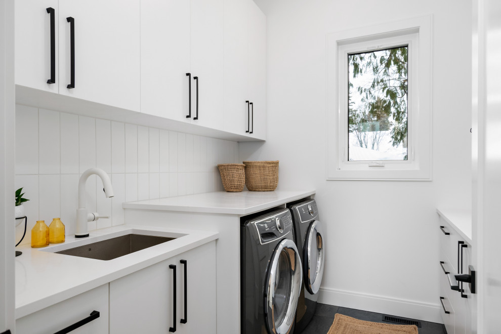 Inredning av en modern vita parallell vitt tvättstuga, med en undermonterad diskho, släta luckor, vita skåp, vita väggar, en tvättmaskin och torktumlare bredvid varandra och svart golv