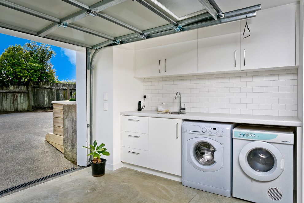 Bild på ett litet funkis linjärt grovkök, med en undermonterad diskho, vita skåp, granitbänkskiva, vita väggar, betonggolv och en tvättmaskin och torktumlare bredvid varandra