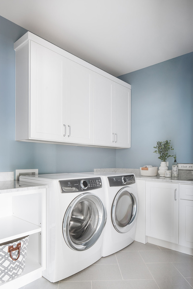 Maritim inredning av en grå grått tvättstuga enbart för tvätt, med skåp i shakerstil, vita skåp, marmorbänkskiva, blå väggar, en tvättmaskin och torktumlare bredvid varandra och grått golv