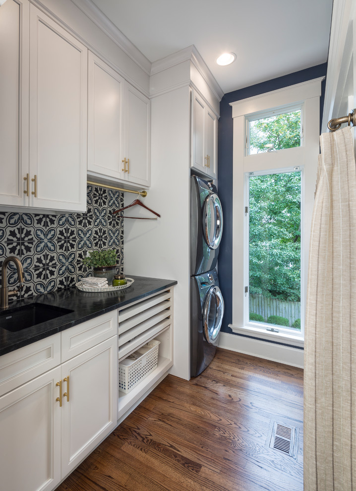 Inspiration for a transitional single-wall dark wood floor and brown floor dedicated laundry room remodel in Cincinnati with an undermount sink, recessed-panel cabinets, white cabinets, blue walls, a stacked washer/dryer and black countertops
