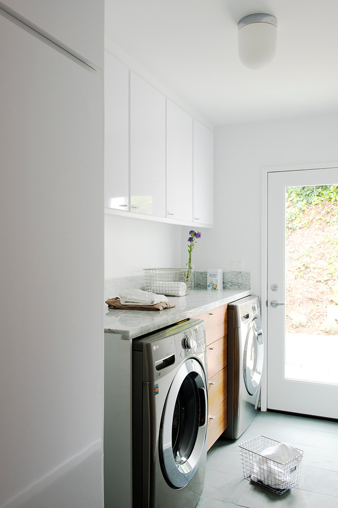 Inredning av en modern mellanstor linjär tvättstuga enbart för tvätt, med släta luckor, marmorbänkskiva, vita väggar, skiffergolv, en tvättmaskin och torktumlare bredvid varandra och vita skåp