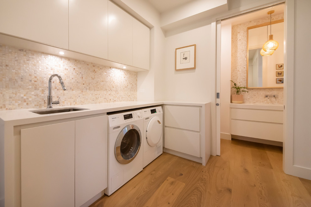 Inspiration for an expansive l-shaped separated utility room in Melbourne with a submerged sink, raised-panel cabinets, white cabinets, engineered stone countertops, white walls, medium hardwood flooring, a side by side washer and dryer and white worktops.