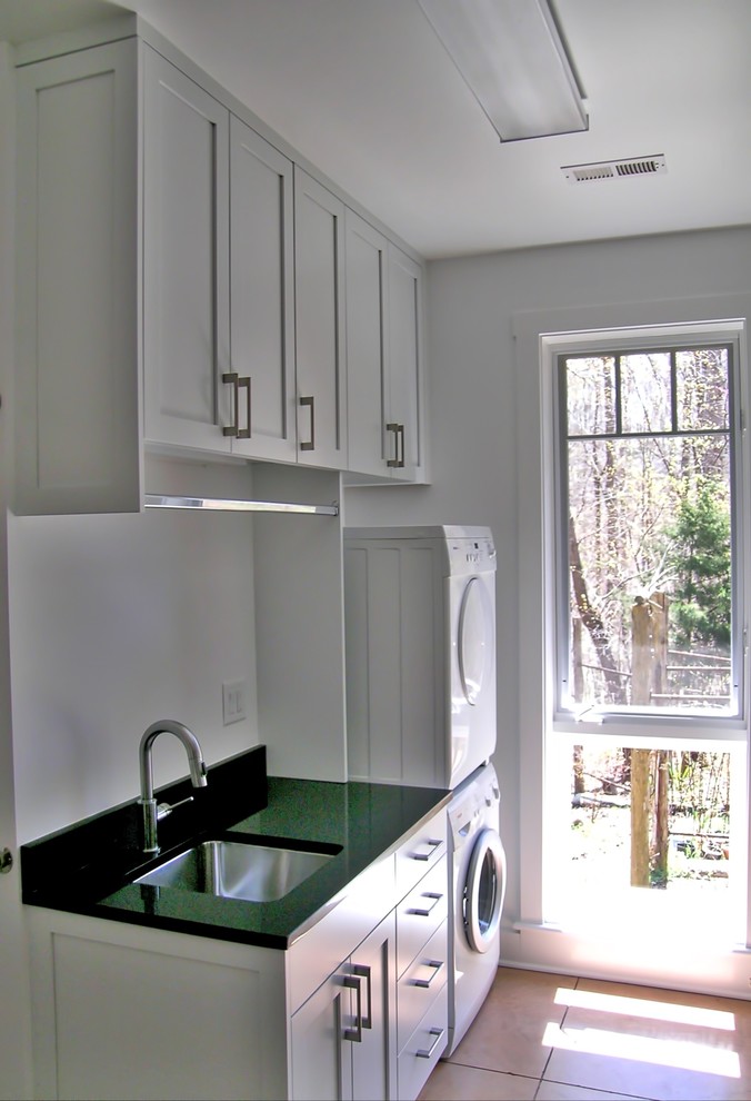 Photo of a small traditional single-wall separated utility room in Raleigh with a submerged sink, shaker cabinets, white cabinets, granite worktops, white walls, porcelain flooring, a stacked washer and dryer and brown floors.
