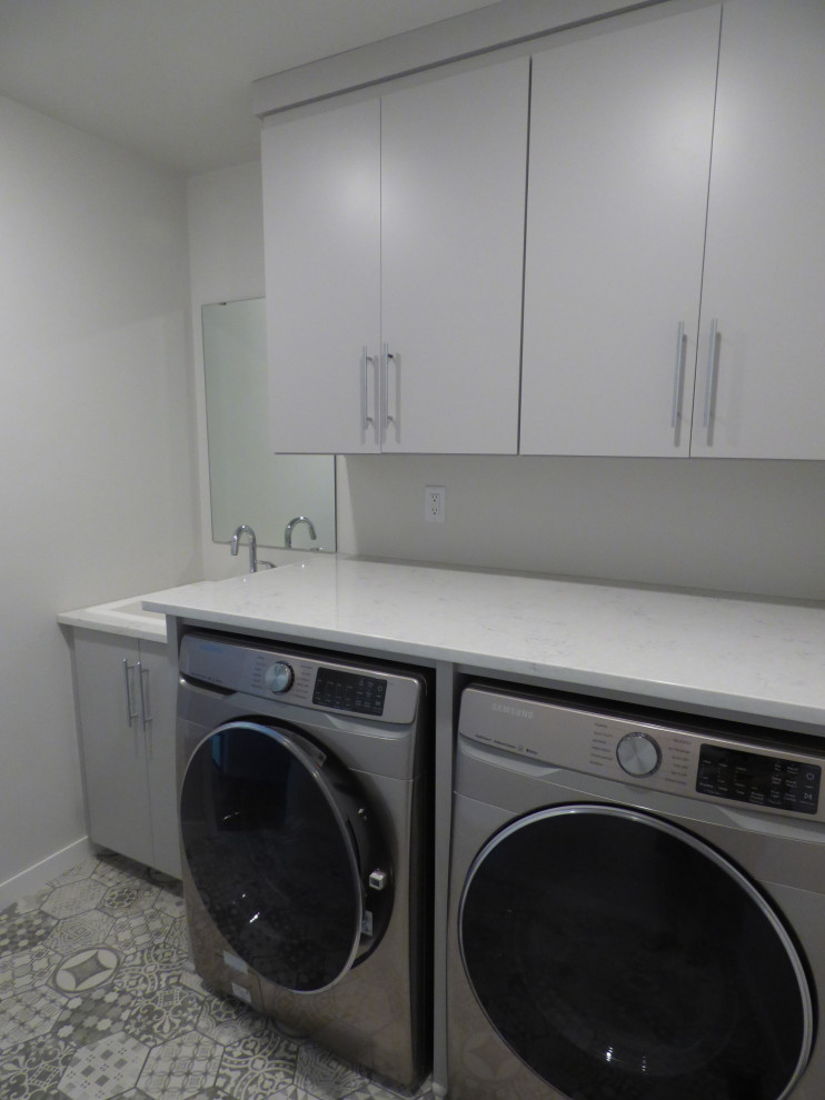 Immagine di una sala lavanderia design di medie dimensioni con lavatoio, ante grigie, top in granito, pareti bianche, pavimento in vinile, lavasciuga, pavimento grigio e top bianco