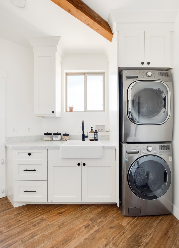 Inspiration for a small rural single-wall separated utility room in Salt Lake City with a belfast sink, shaker cabinets, white cabinets, marble worktops, white walls, porcelain flooring, a stacked washer and dryer, brown floors and grey worktops.