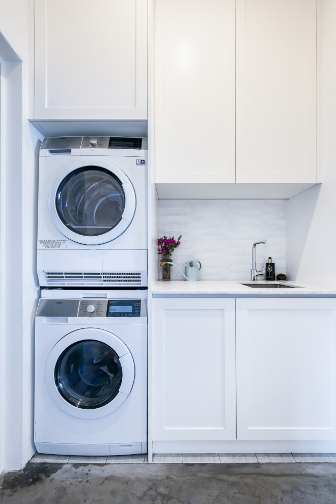 シドニーにあるお手頃価格の小さなビーチスタイルのおしゃれな洗濯室 (I型、アンダーカウンターシンク、シェーカースタイル扉のキャビネット、白いキャビネット、珪岩カウンター、白いキッチンパネル、セラミックタイルのキッチンパネル、白い壁、上下配置の洗濯機・乾燥機、グレーのキッチンカウンター) の写真