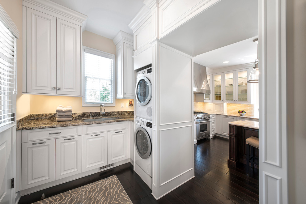 Elegant l-shaped dark wood floor laundry room photo in Detroit with an undermount sink, raised-panel cabinets, white cabinets, beige walls and a stacked washer/dryer