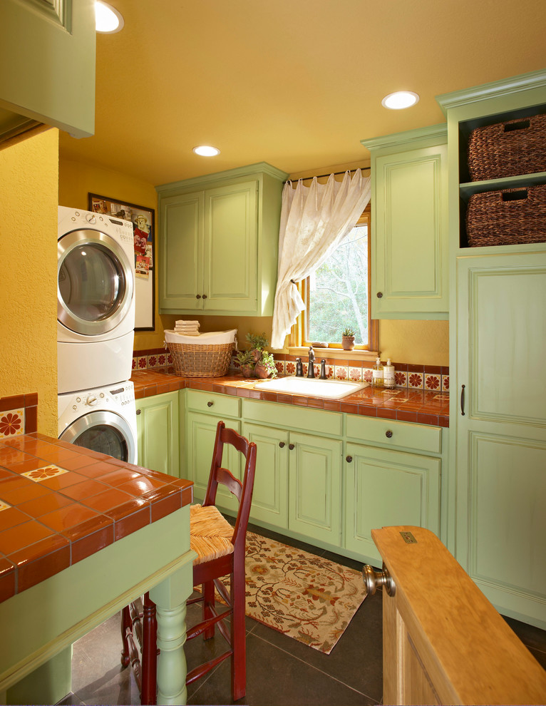 ロサンゼルスにあるお手頃価格の小さなトラディショナルスタイルのおしゃれな洗濯室 (レイズドパネル扉のキャビネット、緑のキャビネット、タイルカウンター、マルチカラーのキッチンパネル、黄色い壁、ドロップインシンク、磁器タイルの床、上下配置の洗濯機・乾燥機、黒い床、赤いキッチンカウンター、コの字型) の写真