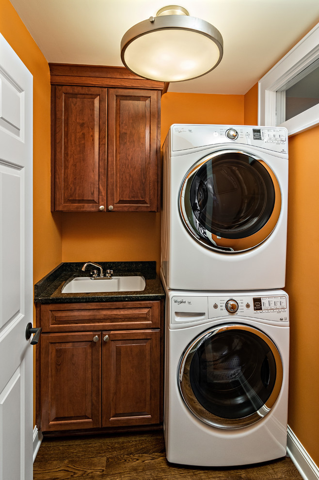 Einzeilige, Kleine Rustikale Waschküche mit Unterbauwaschbecken, profilierten Schrankfronten, dunklen Holzschränken, Granit-Arbeitsplatte, oranger Wandfarbe, braunem Holzboden, Waschmaschine und Trockner gestapelt, braunem Boden und schwarzer Arbeitsplatte in Minneapolis
