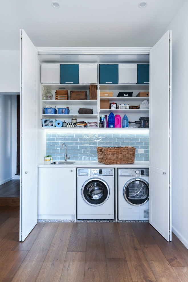 Einzeiliger Moderner Hauswirtschaftsraum mit Waschmaschinenschrank, Unterbauwaschbecken, flächenbündigen Schrankfronten, weißen Schränken, weißer Wandfarbe, dunklem Holzboden, Waschmaschine und Trockner nebeneinander, braunem Boden und weißer Arbeitsplatte in Sydney