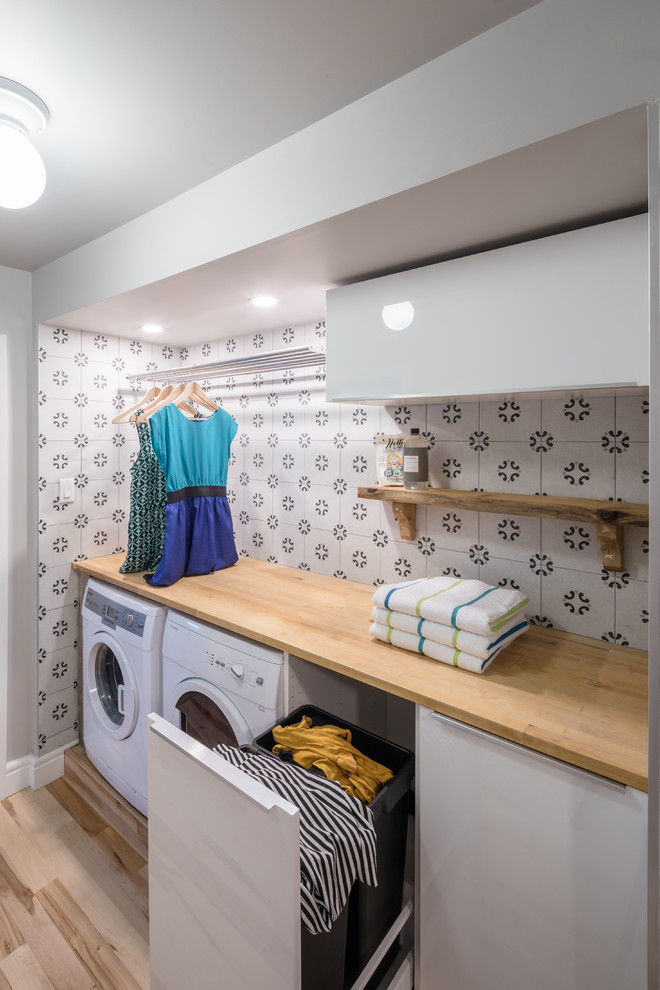 Ejemplo de cuarto de lavado lineal actual pequeño con armarios con paneles lisos, puertas de armario blancas, encimera de madera, paredes grises, suelo de madera clara, lavadora y secadora juntas y encimeras beige