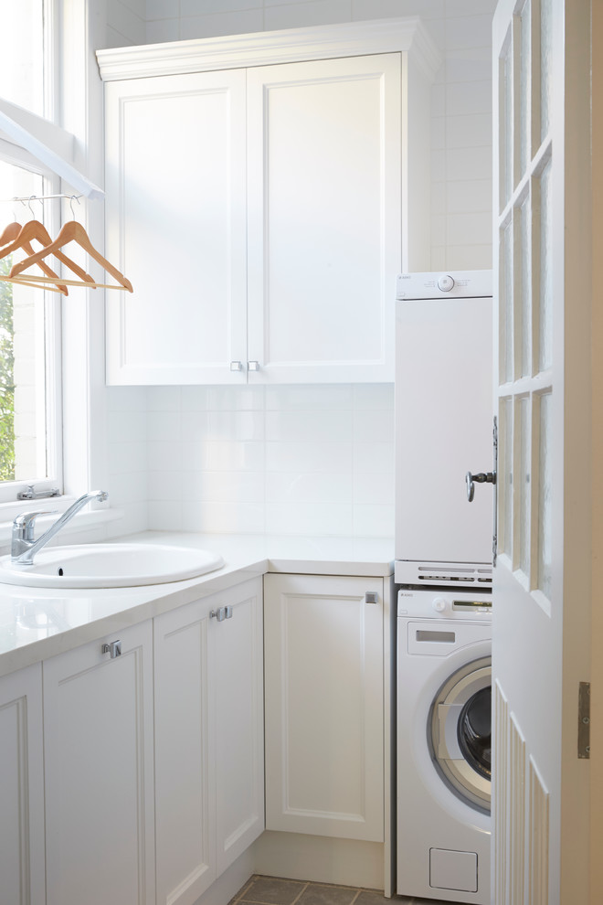 Example of a mid-sized classic l-shaped dedicated laundry room design in Sydney with a drop-in sink, a stacked washer/dryer, recessed-panel cabinets, white cabinets, white walls and white countertops