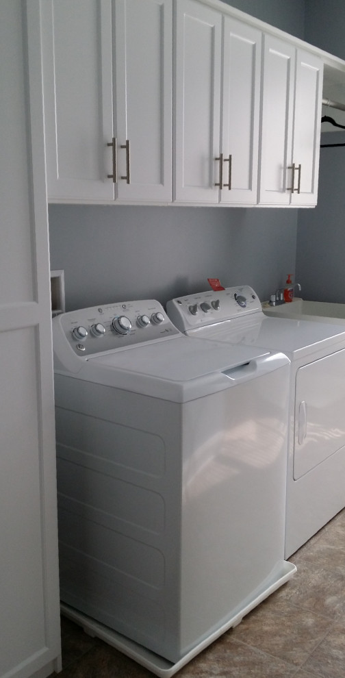 Mid-sized single-wall ceramic tile dedicated laundry room photo with gray walls, a side-by-side washer/dryer, an utility sink, recessed-panel cabinets and white cabinets
