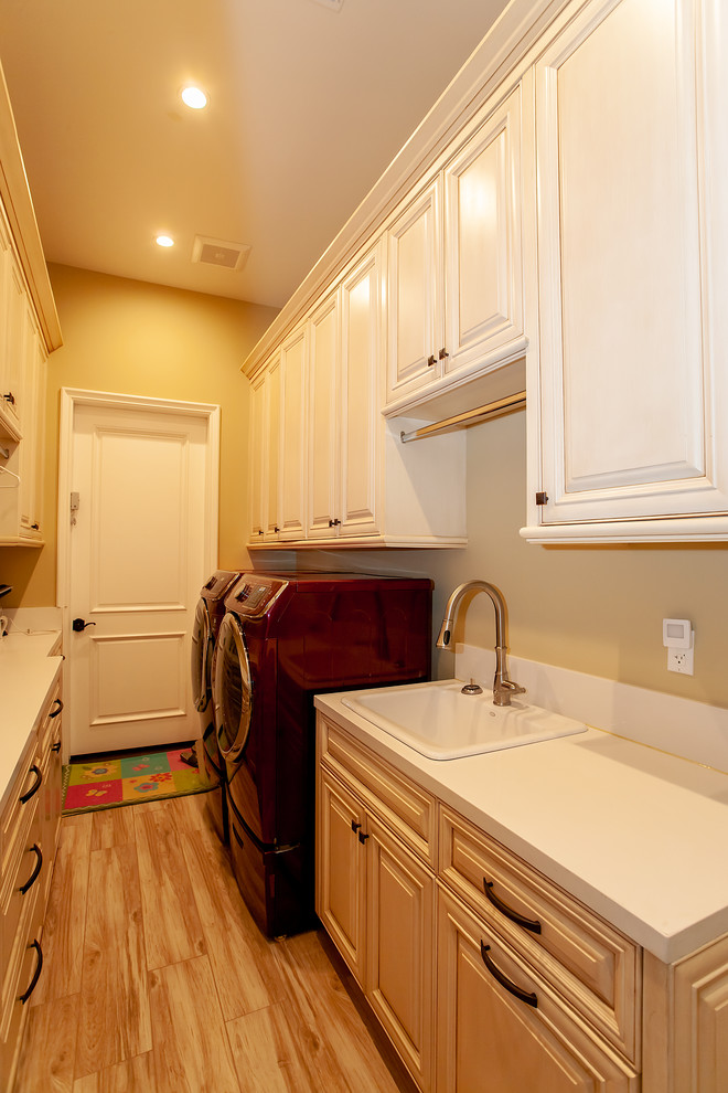Example of a mid-sized tuscan galley light wood floor and brown floor dedicated laundry room design in Los Angeles with a single-bowl sink, raised-panel cabinets, beige cabinets, solid surface countertops, beige walls, a side-by-side washer/dryer and white countertops