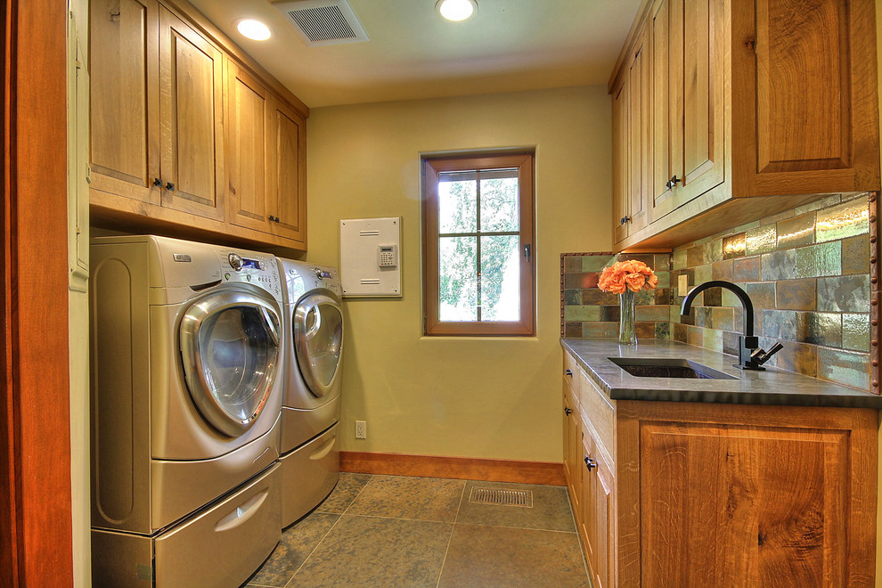 Immagine di una sala lavanderia stile americano con lavello sottopiano, ante con bugna sagomata, ante in legno scuro, top in superficie solida, pavimento in ardesia e lavatrice e asciugatrice affiancate