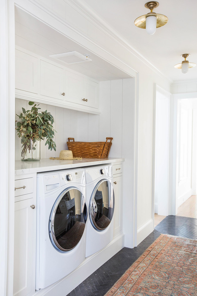 Imagen de armario lavadero lineal campestre con armarios estilo shaker, puertas de armario blancas, encimera de mármol, lavadora y secadora juntas, suelo negro y encimeras blancas