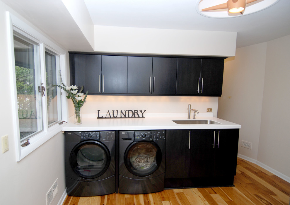 Small single-wall separated utility room in Milwaukee with a submerged sink, flat-panel cabinets, composite countertops, white walls, medium hardwood flooring, a side by side washer and dryer and black cabinets.