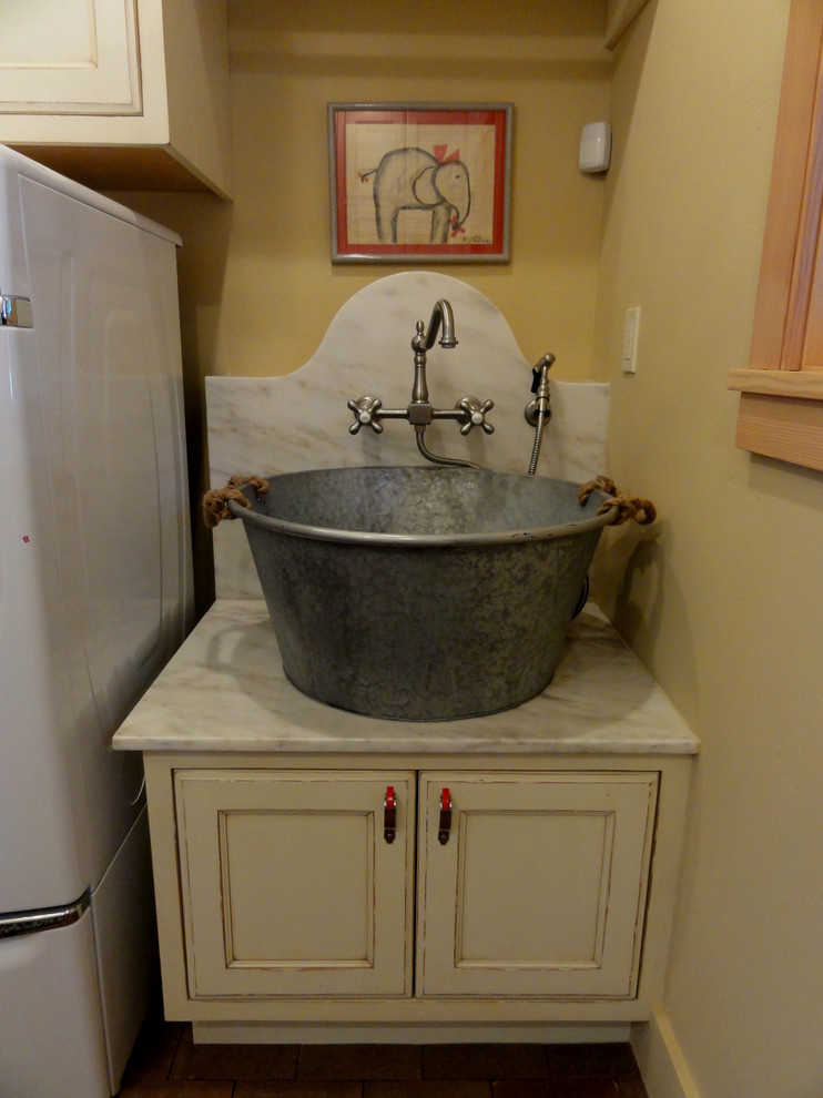 Foto på en mellanstor lantlig tvättstuga, med skåp i slitet trä, marmorbänkskiva, en tvättmaskin och torktumlare bredvid varandra och luckor med infälld panel