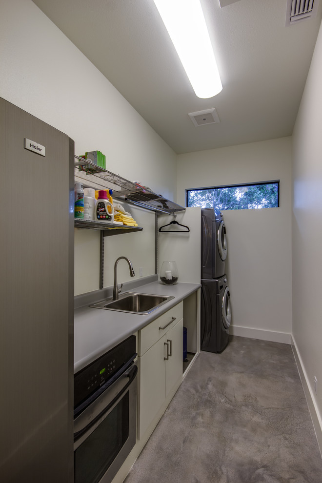 Design ideas for a medium sized galley utility room with a built-in sink, flat-panel cabinets, white cabinets, laminate countertops, grey walls, concrete flooring, a stacked washer and dryer and grey floors.