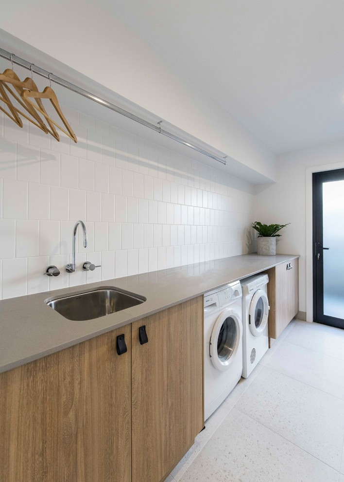 Einzeilige Moderne Waschküche mit Unterbauwaschbecken, hellbraunen Holzschränken, weißer Wandfarbe, Keramikboden, Waschmaschine und Trockner nebeneinander, flächenbündigen Schrankfronten, grauem Boden und beiger Arbeitsplatte in Brisbane