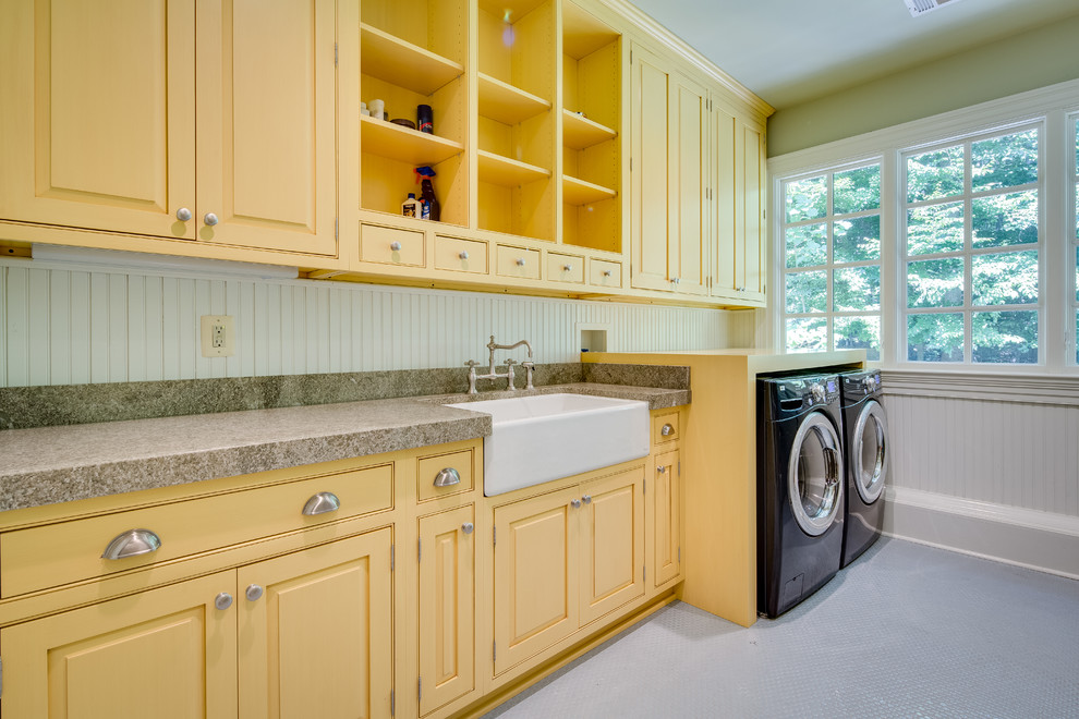 Bild på en stor lantlig grå linjär grått tvättstuga enbart för tvätt, med en rustik diskho, granitbänkskiva, gröna väggar, klinkergolv i keramik, en tvättmaskin och torktumlare bredvid varandra, gula skåp och luckor med profilerade fronter