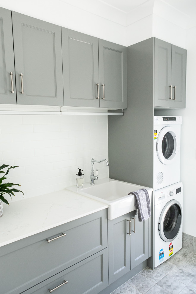 This is an example of a rural separated utility room in Sunshine Coast with a belfast sink, recessed-panel cabinets, grey cabinets, engineered stone countertops, white walls, ceramic flooring, a stacked washer and dryer, grey floors and white worktops.
