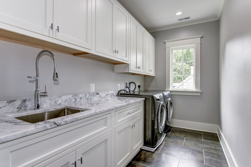 Exempel på en stor amerikansk linjär tvättstuga enbart för tvätt, med en undermonterad diskho, luckor med infälld panel, vita skåp, marmorbänkskiva, grå väggar, klinkergolv i keramik, en tvättmaskin och torktumlare bredvid varandra och brunt golv