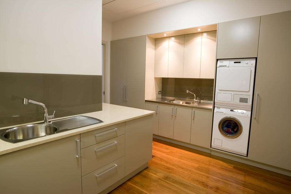 Photo of a large modern separated utility room in Sydney with a built-in sink, flat-panel cabinets, engineered stone countertops, medium hardwood flooring, a stacked washer and dryer, beige cabinets and white walls.