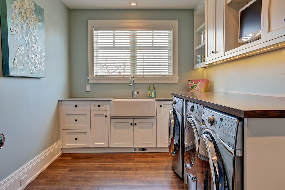 Klassisk inredning av en mellanstor l-formad tvättstuga enbart för tvätt, med en rustik diskho, skåp i shakerstil, vita skåp, träbänkskiva, mellanmörkt trägolv, en tvättmaskin och torktumlare bredvid varandra och grå väggar