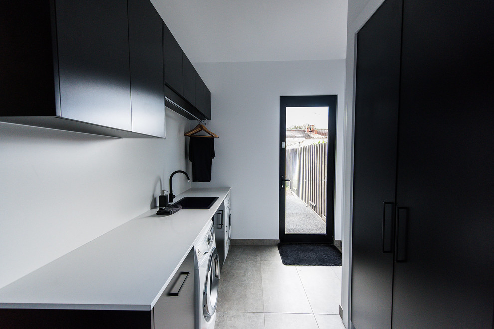 Bild på en mellanstor funkis tvättstuga, med en nedsänkt diskho, svarta skåp, bänkskiva i kvarts, vita väggar, betonggolv, en tvättmaskin och torktumlare bredvid varandra och grått golv
