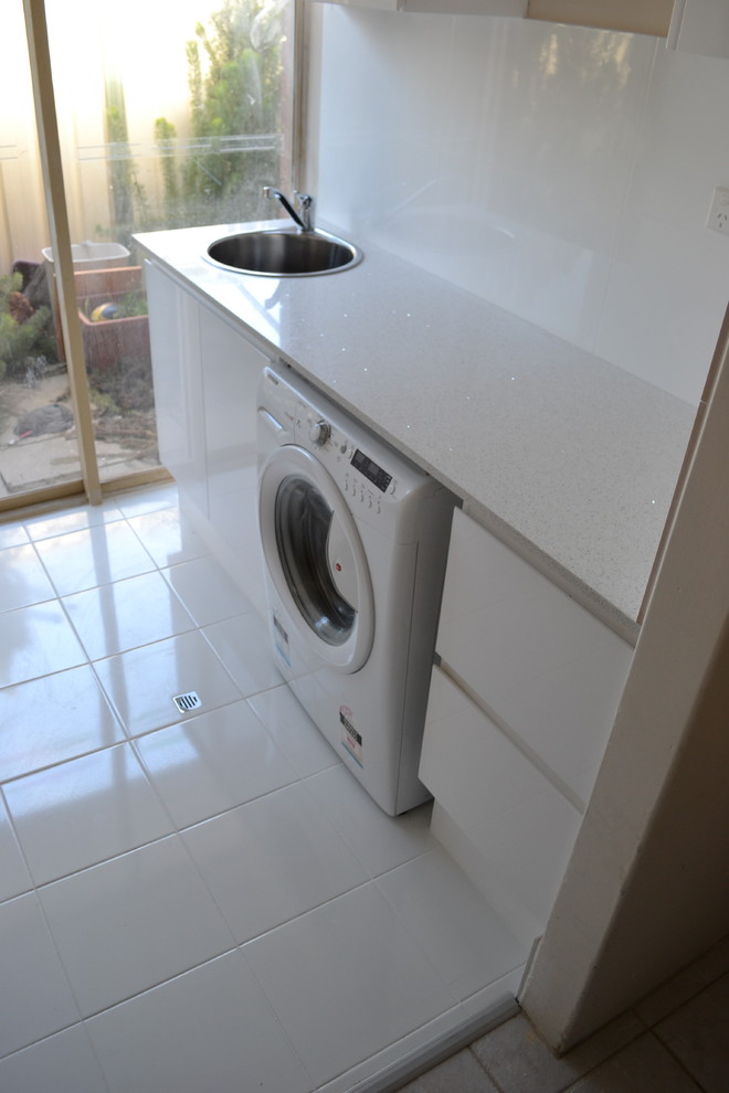 Idéer för små funkis linjära tvättstugor enbart för tvätt, med en nedsänkt diskho, luckor med lamellpanel, vita skåp, bänkskiva i kvarts, vita väggar, klinkergolv i porslin, en tvättmaskin och torktumlare bredvid varandra och vitt golv