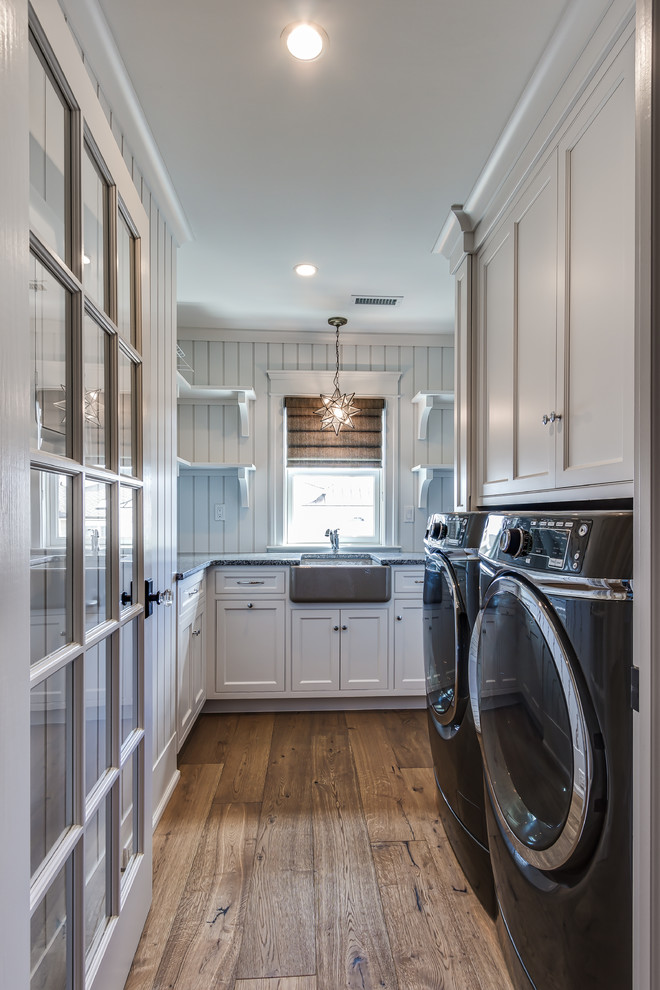 Inspiration for a large coastal l-shaped medium tone wood floor laundry room remodel in Philadelphia with a farmhouse sink, flat-panel cabinets, white cabinets, granite countertops, white walls and a side-by-side washer/dryer