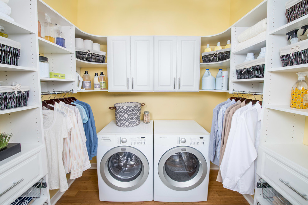 Exempel på en stor klassisk u-formad tvättstuga enbart för tvätt, med luckor med upphöjd panel, vita skåp, gula väggar, mellanmörkt trägolv, en tvättmaskin och torktumlare bredvid varandra och brunt golv