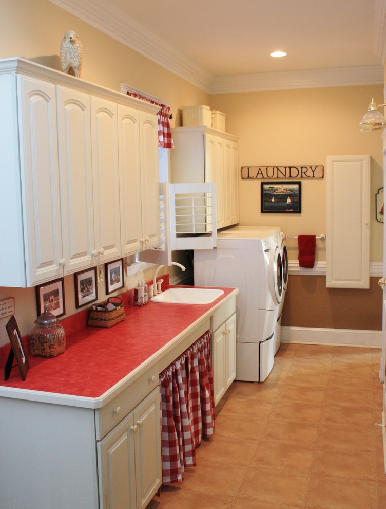 ワシントンD.C.にある広いカントリー風のおしゃれな洗濯室 (レイズドパネル扉のキャビネット、白いキャビネット、ベージュの壁、左右配置の洗濯機・乾燥機、I型、ドロップインシンク、ラミネートの床、赤いキッチンカウンター) の写真