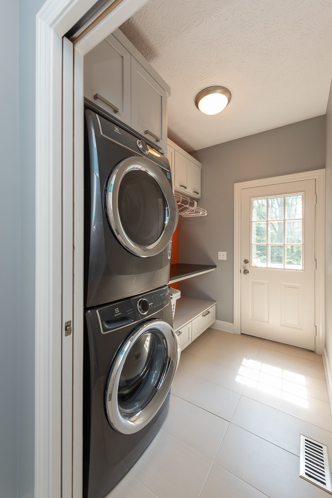 Inspiration for a small traditional single-wall utility room in Indianapolis with recessed-panel cabinets, grey cabinets, granite worktops, grey walls, ceramic flooring, a stacked washer and dryer, white floors and black worktops.